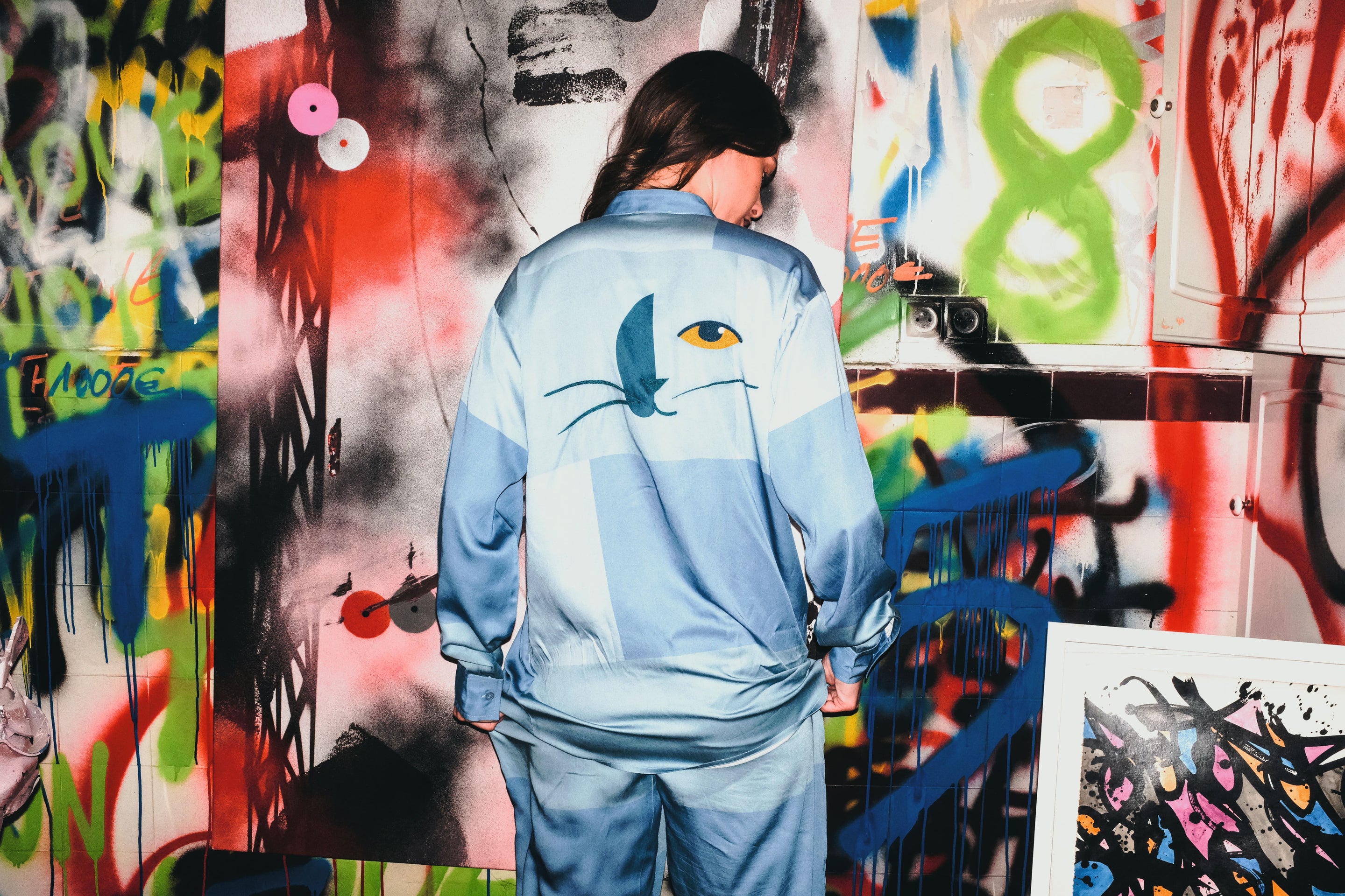 Person wearing a blue tracksuit with a cat design in front of a colorful graffiti wall.