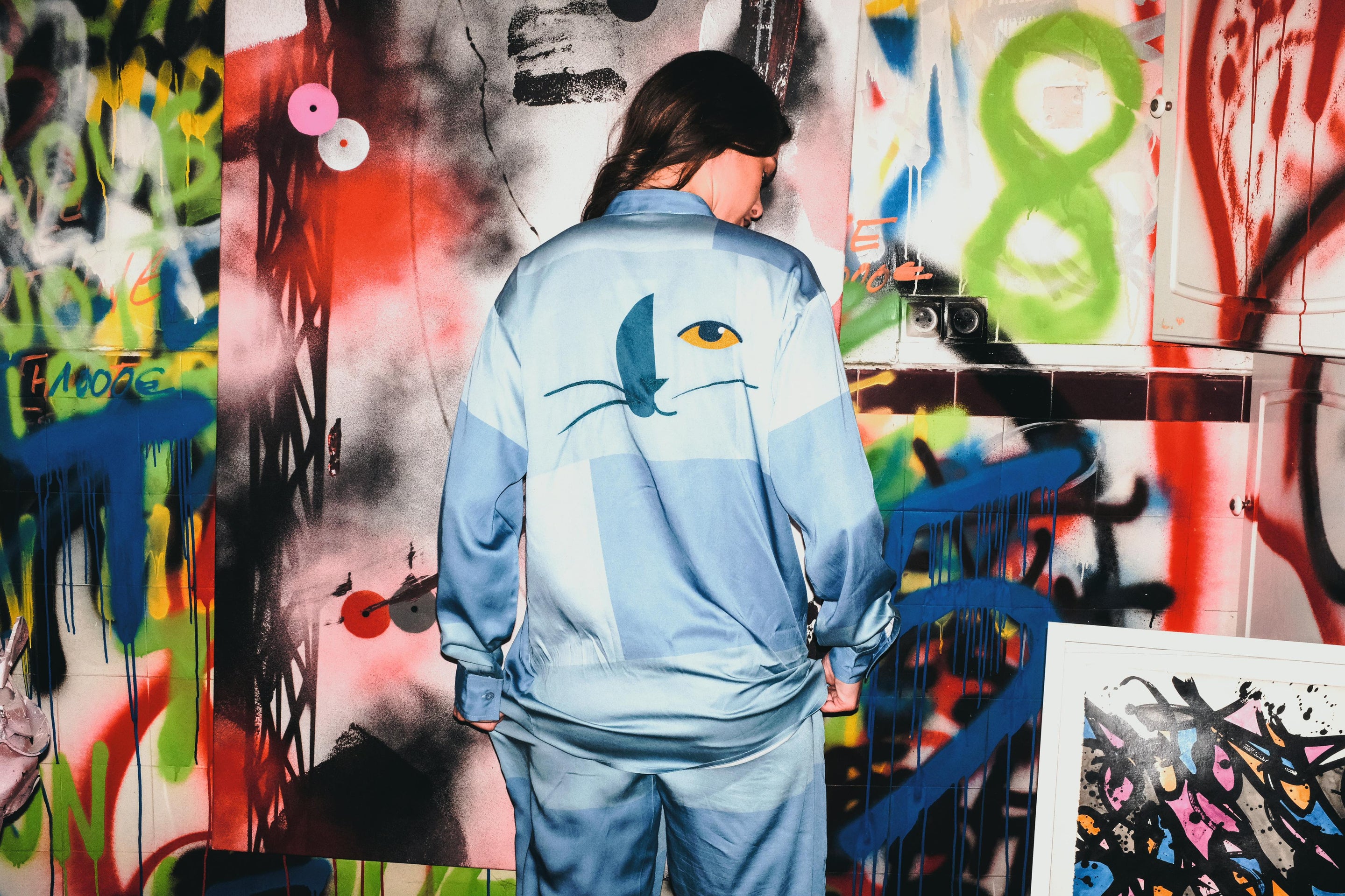 Person wearing a blue tracksuit with a cat design in front of a colorful graffiti wall.